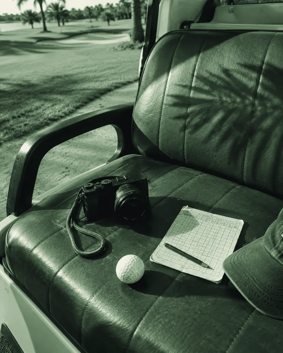 A camera, a golf ball, a pencilled scorecard and a cap on the green vinyl seat of a cart, palm shadow across it